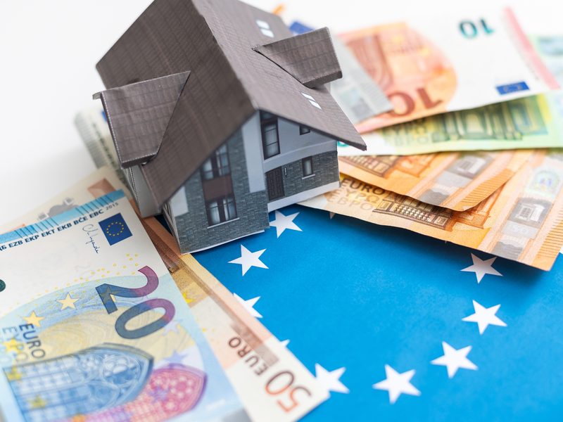 Una maqueta de casa con el tejado gris sobre billetes de euro y una bandera azul con estrellas