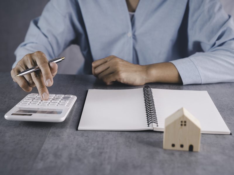 Una persona hace cuentas con una calculadora junto a una libreta en blanco y una casita de madera, sobre una mesa gris
