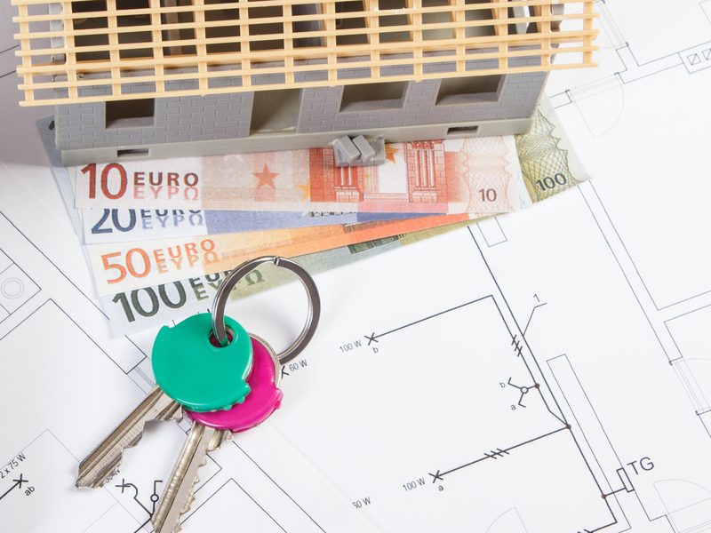 Maqueta de una casa en obras sobre billetes de euro, junto a un llavero con dos llaves encima de unos planos de instalación eléctrica