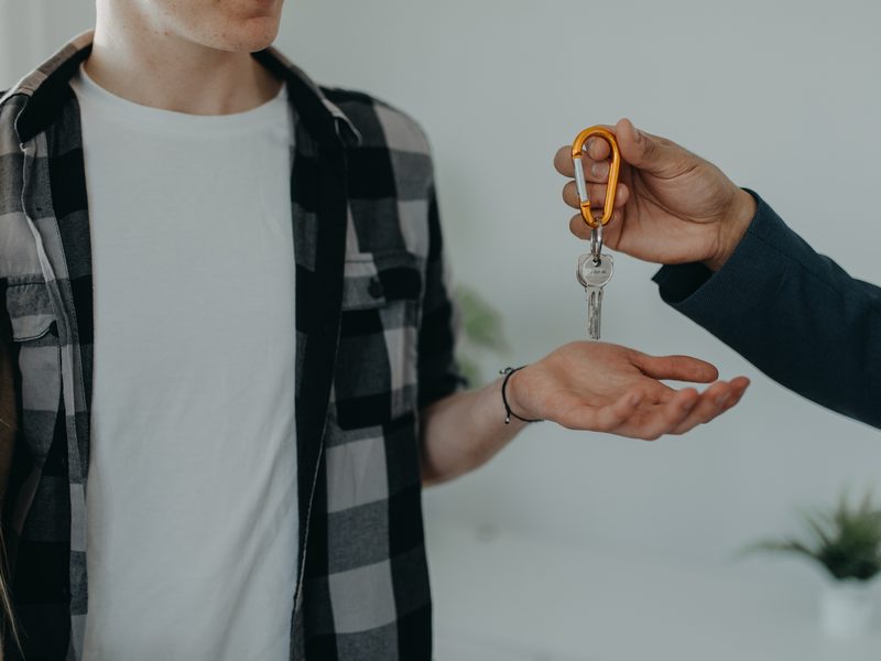 Un joven con camisa de cuadros tiende la mano abierta y otra persona le entrega unas llaves con un llavero naranja