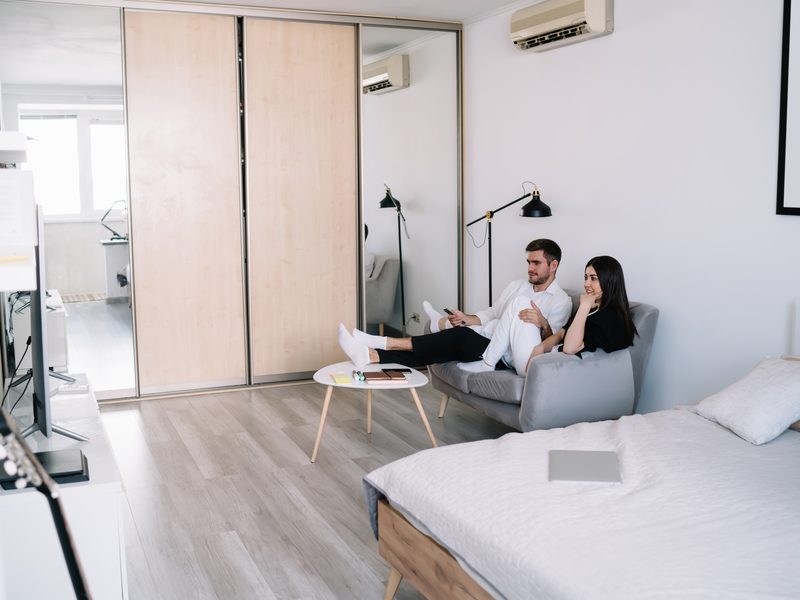 Una pareja descansa en el sofá de un estudio luminoso, con la cama en primer plano y un armario de puertas de madera al fondo