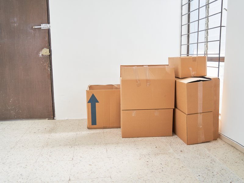 El inquilino que deja el piso antes de que acabe el contrato Cajas de cartón cerradas con cinta y apiladas junto a la puerta de una habitación vacía con suelo de terrazo y una ventana con reja