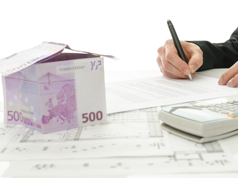 Una mano firma un documento junto a una calculadora y a una casita hecha con billetes de 500 euros, sobre unos planos