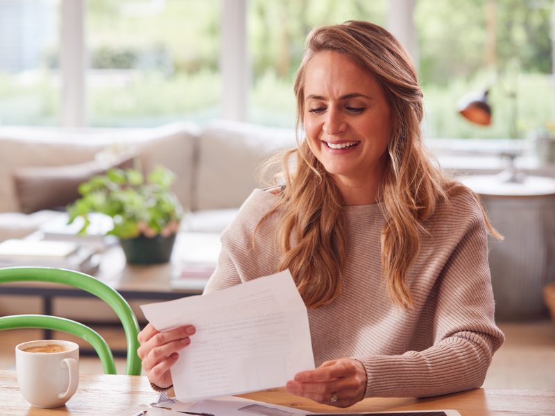 Mujer sonriente leyendo una carta en la mesa del salón de su casa, junto a un café