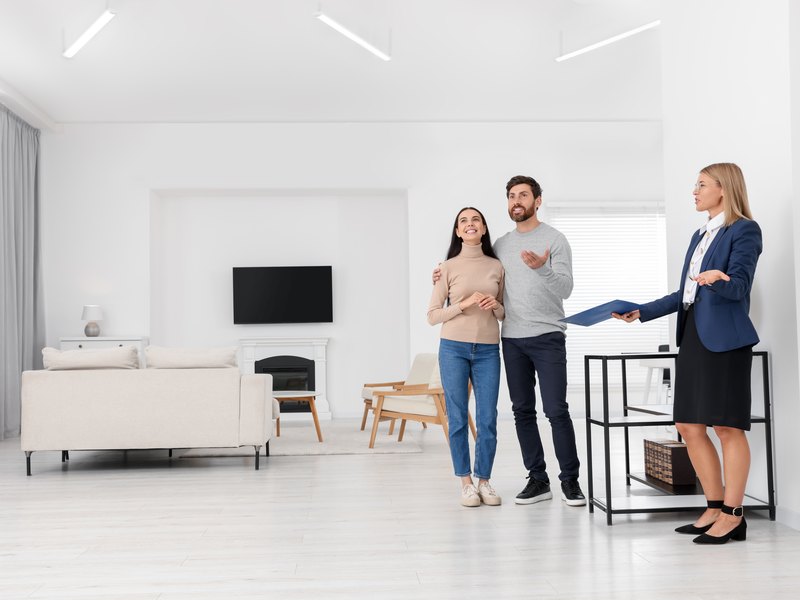 Agente inmobiliaria con una carpeta enseñando un salón blanco y luminoso a una pareja que visita el piso