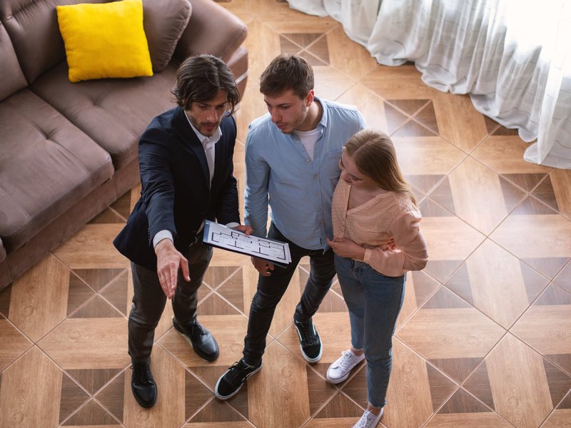 Visita al piso con el agente inmobiliario, alquilar tu piso por inmobiliaria en Zaragoza Inmobiliaria en Zaragoza para el alquiler: un agente con el plano del piso enseña el salón a una pareja joven