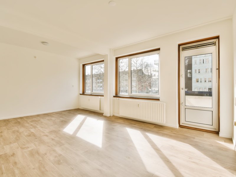 Salón vacío y soleado de un piso sin muebles, con suelo de madera clara, radiadores bajo las ventanas y una puerta al balcón