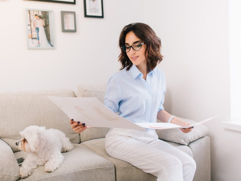 Una mujer con gafas revisa unos papeles sentada en el sofá de su casa, con su perro blanco al lado y fotos colgadas en la pared