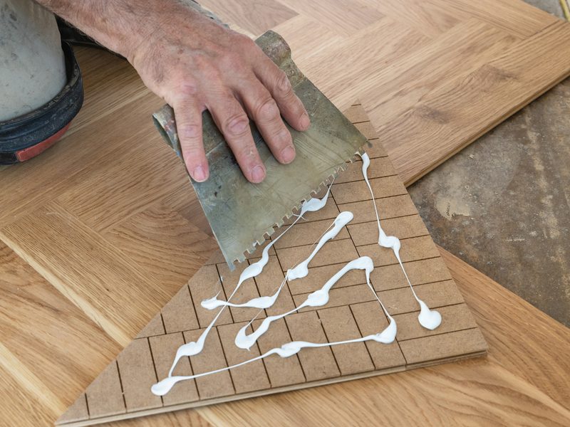 Un operario extiende adhesivo con una llana dentada sobre una pieza de parquet en espiga
