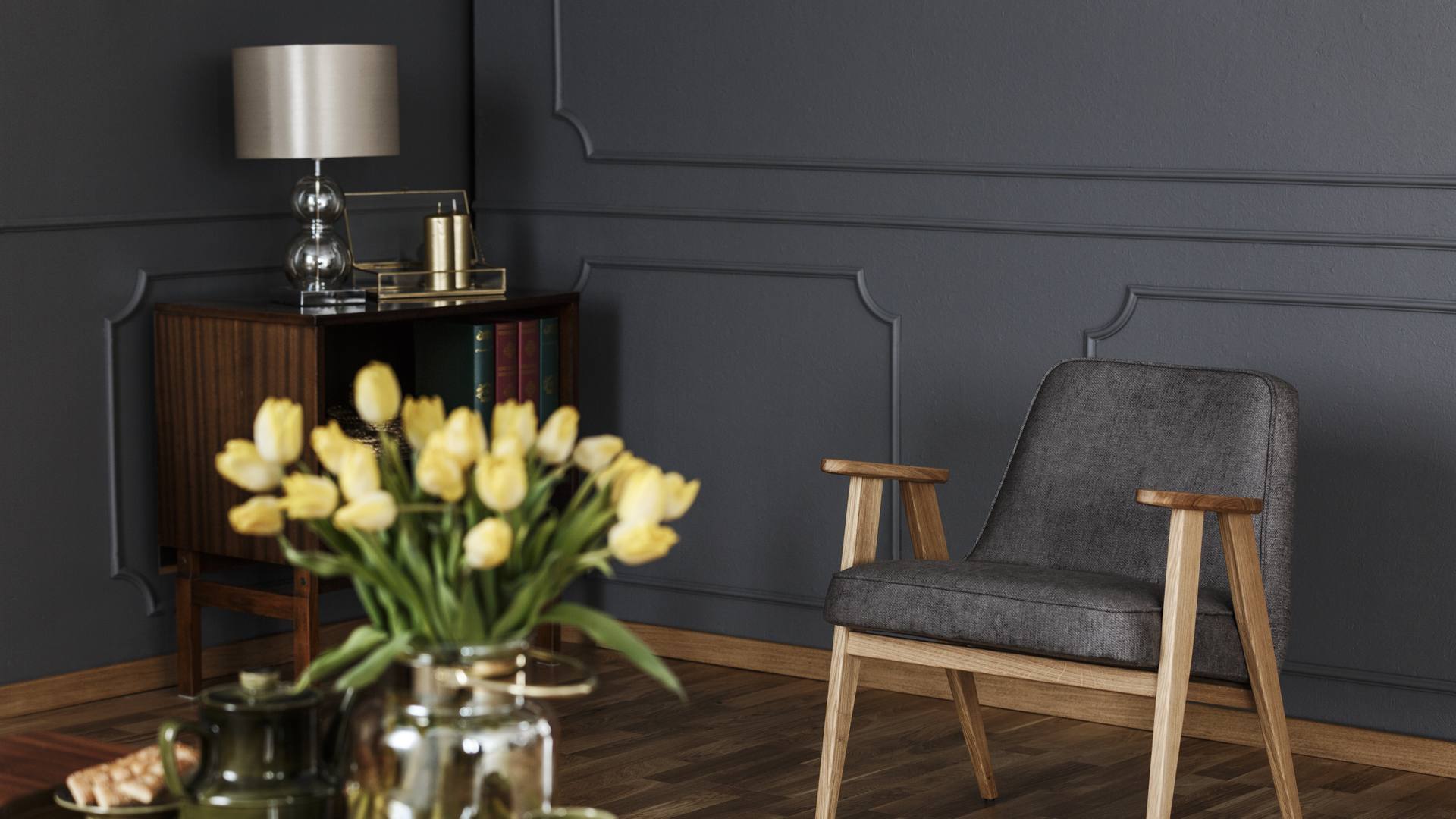 Un sillón de madera tapizado en gris y una mesita con libros y una lámpara, junto a un jarrón de tulipanes amarillos, en el salón de un piso