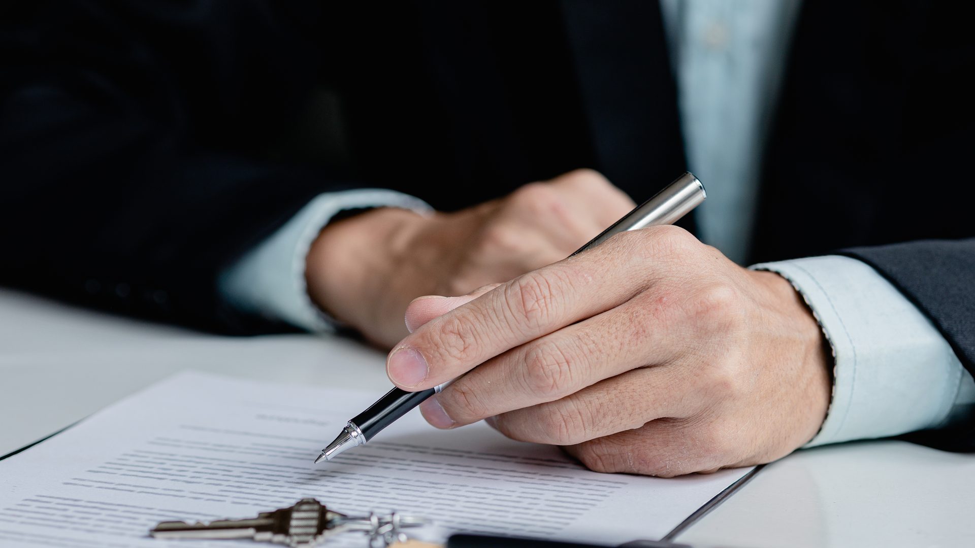 Mano con un bolígrafo a punto de firmar un documento, con la llave de un piso encima del papel