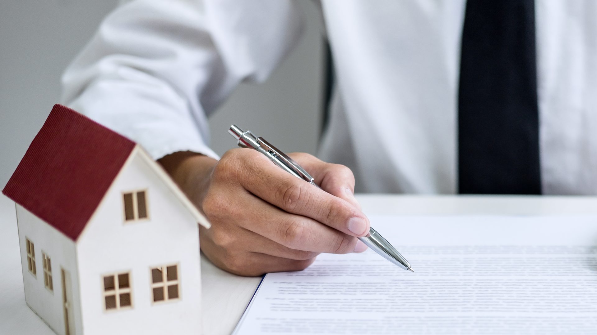 Una mano firma un documento con un bolígrafo, junto a una casita de madera de tejado rojo