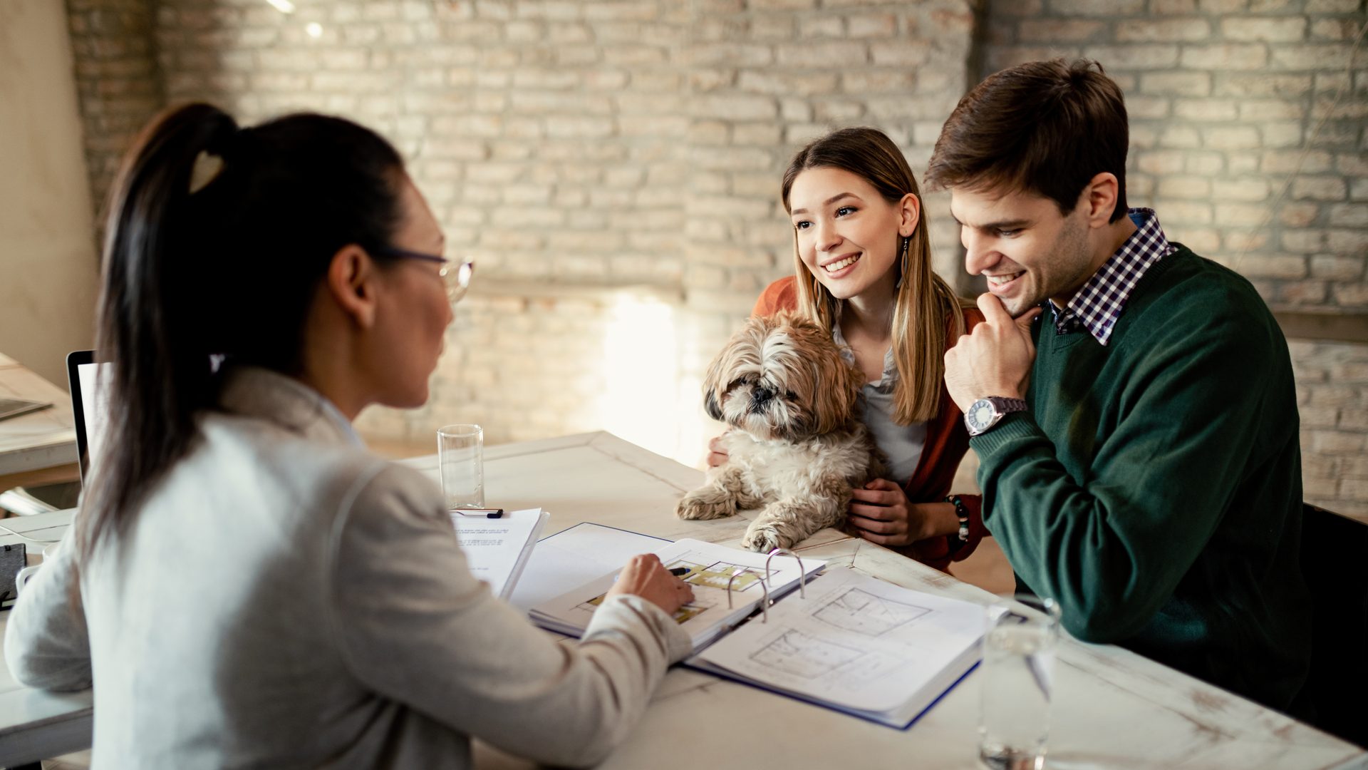 Una mujer con gafas y coleta, de espaldas, enseña los planos de un piso en un archivador a una pareja sonriente que sostiene a un perro pequeño, en una mesa junto a una pared de ladrillo
