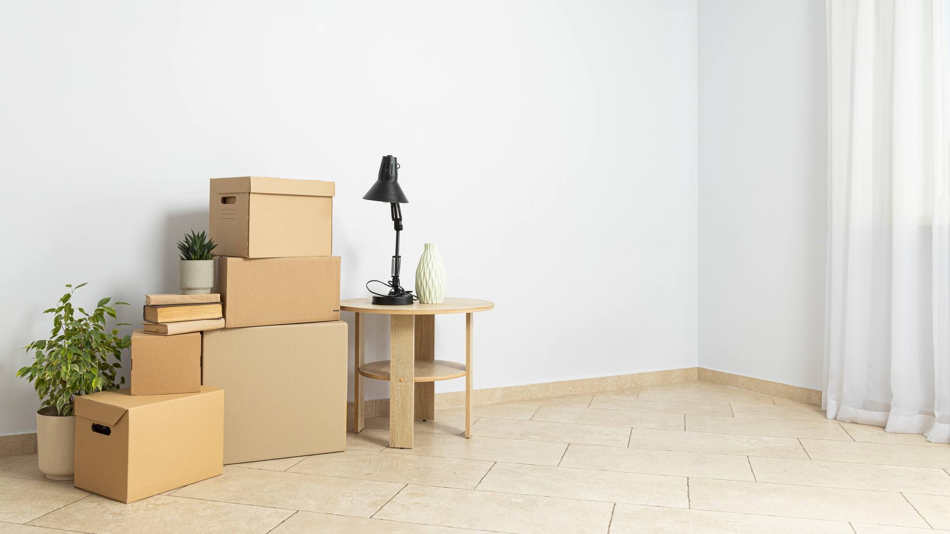 Una habitación vacía de paredes blancas con cajas de cartón apiladas, una mesita con una lámpara y un jarrón, dos plantas y una cortina