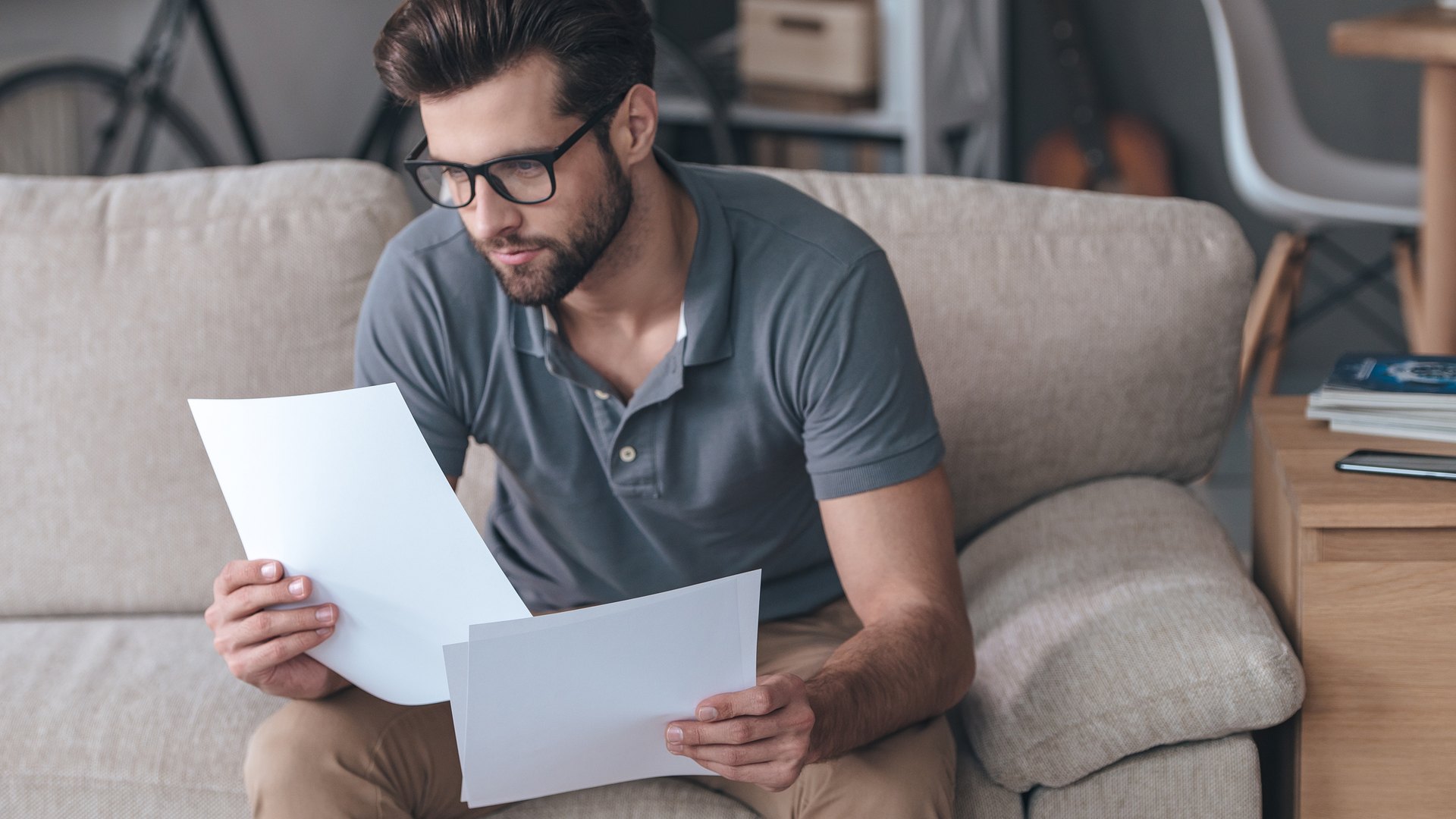 Hombre con gafas sentado en el sofá de su salón leyendo con atención dos hojas de papel
