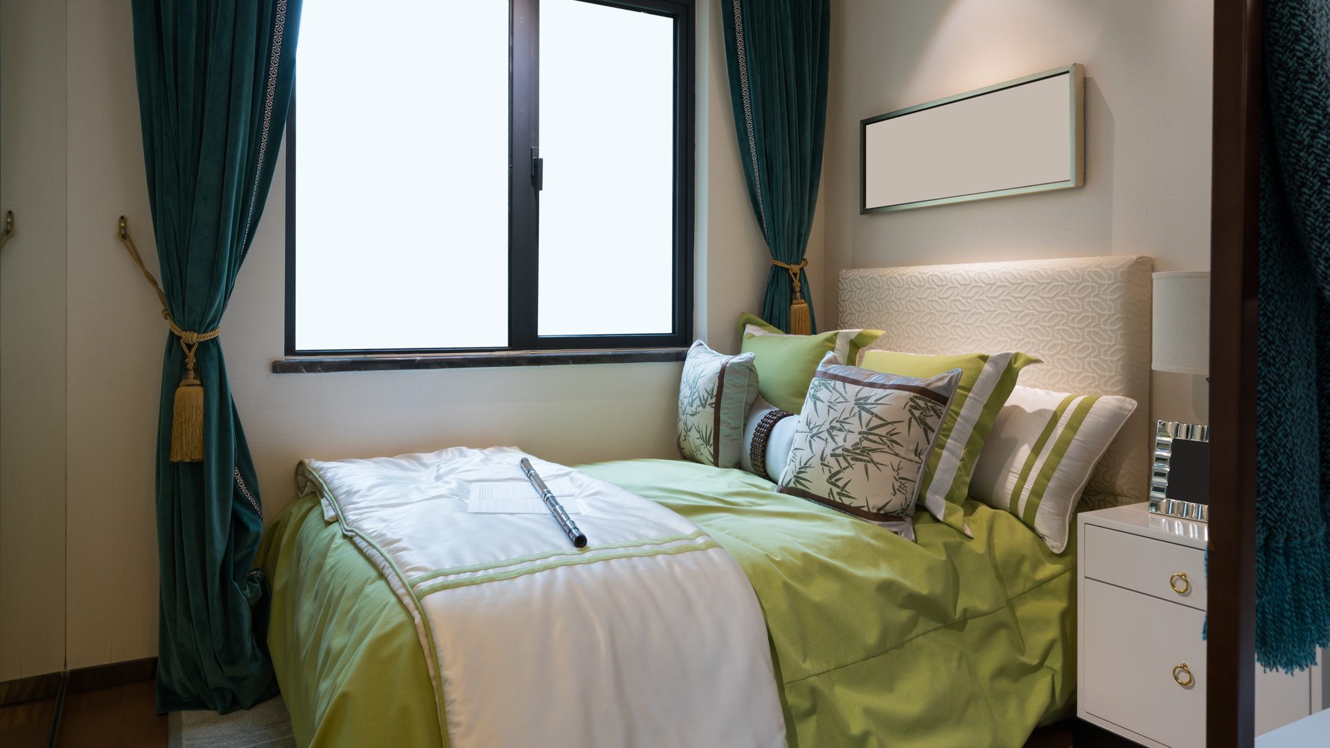 Habitación con una cama de colcha verde y blanca, cojines estampados, una mesilla blanca y cortinas verdes junto a la ventana