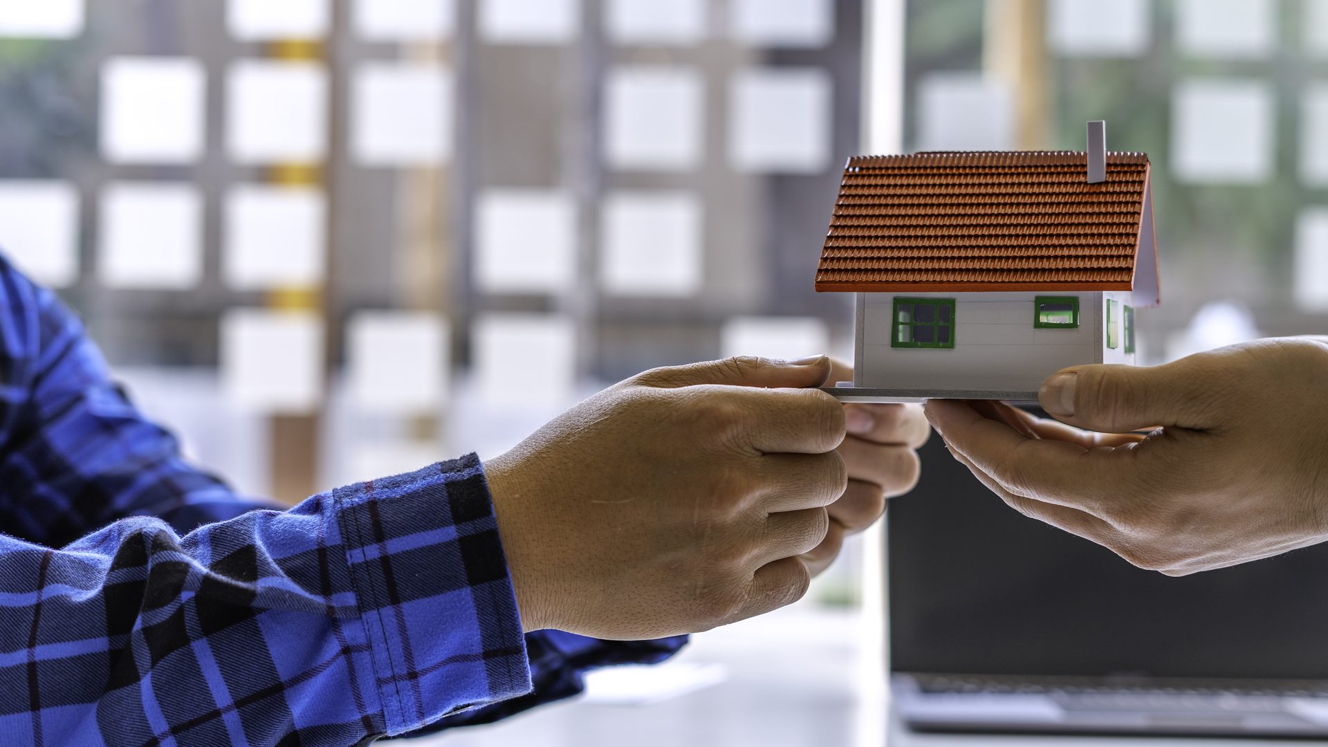 Dos manos se pasan una maqueta de casa con tejado de teja sobre una mesa, con un portátil al fondo