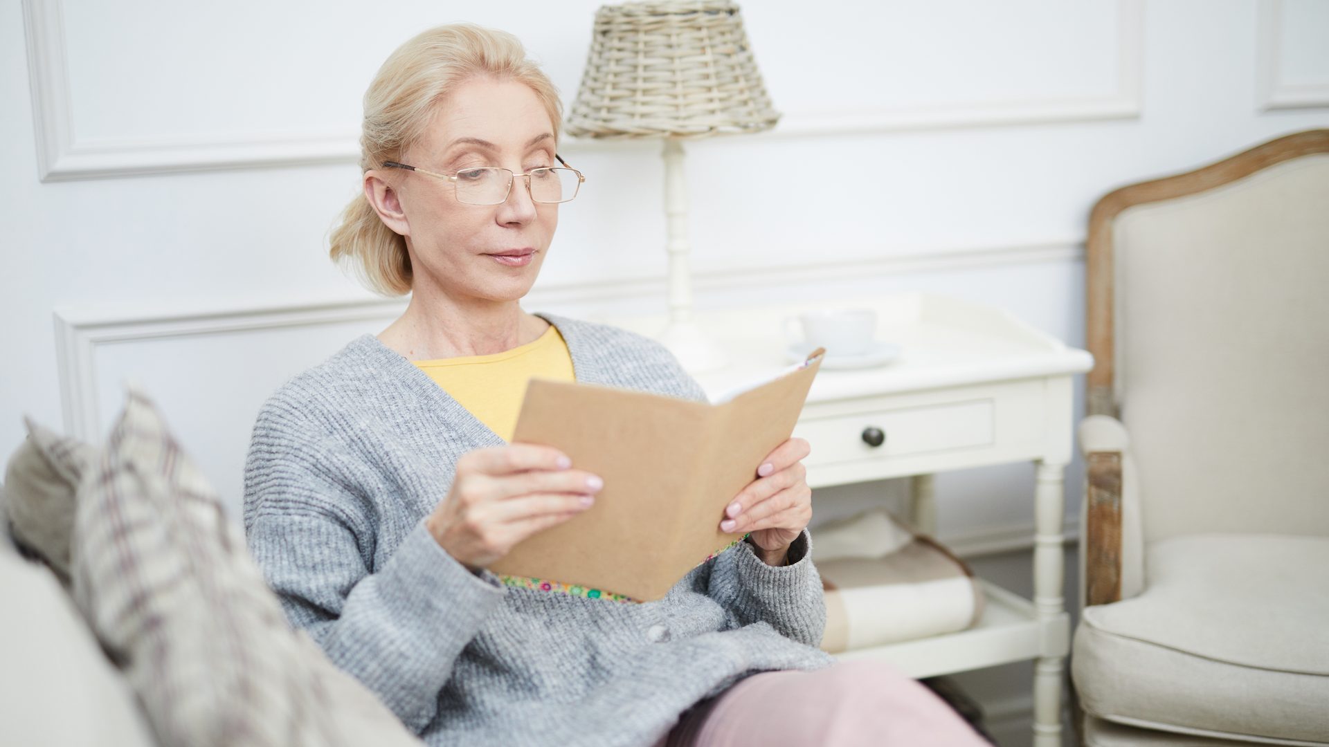 Una mujer con gafas lee un cuaderno sentada en el sofá de su salón, junto a una mesita blanca con una lámpara de mimbre y un sillón tapizado