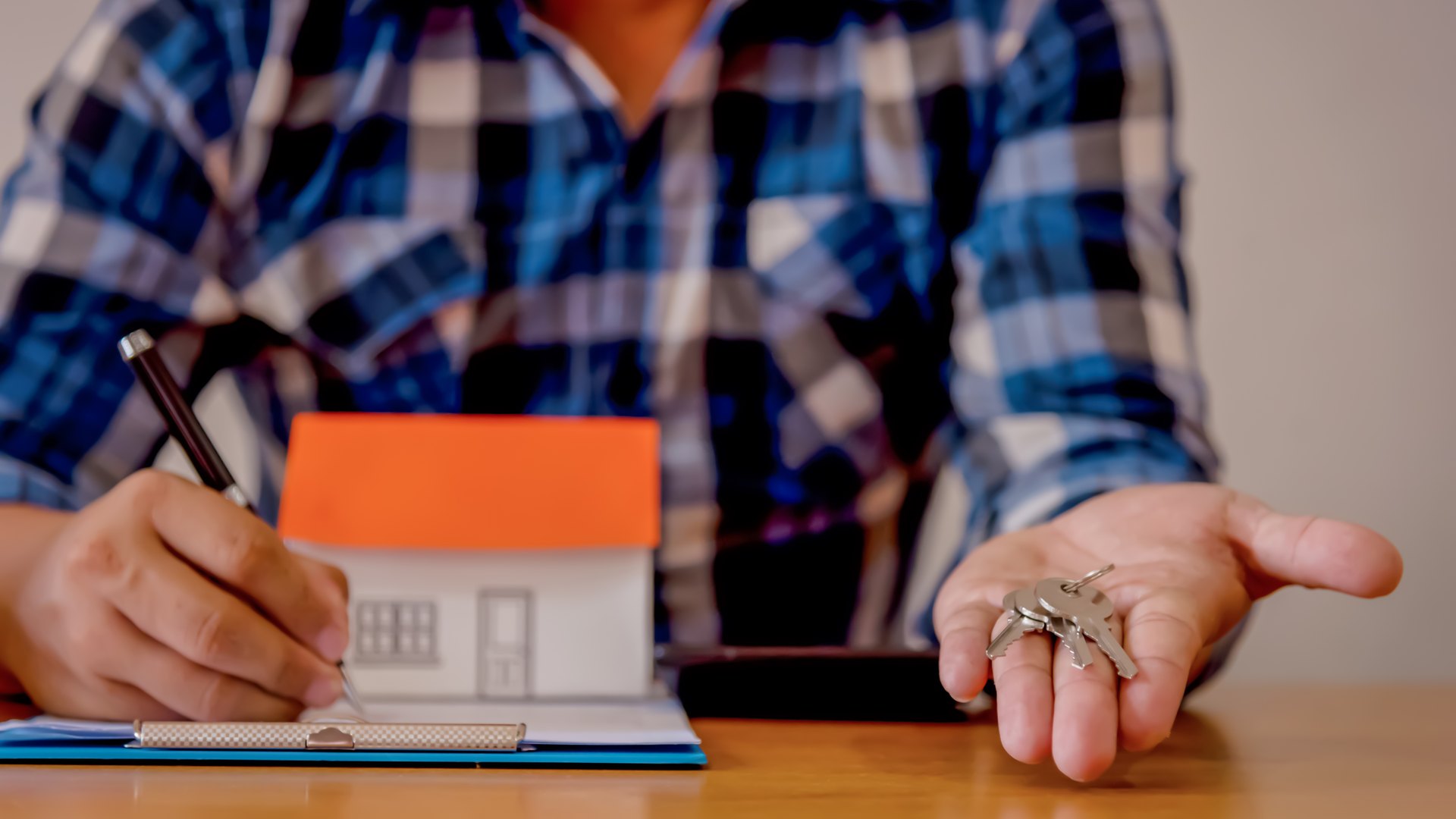 Hombre con camisa de cuadros que firma un contrato en una carpeta junto a una maqueta de casa y muestra en la otra mano las llaves de la vivienda
