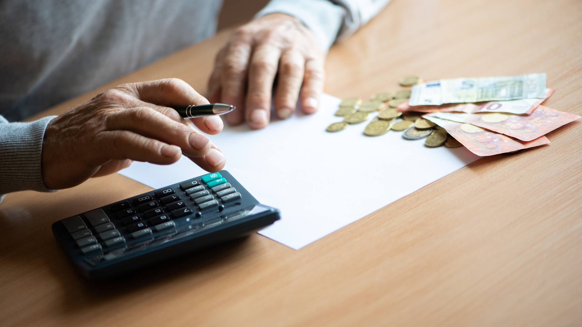 Unas manos con un bolígrafo sobre un papel en blanco, junto a billetes y monedas de euro y una calculadora, en una mesa de madera