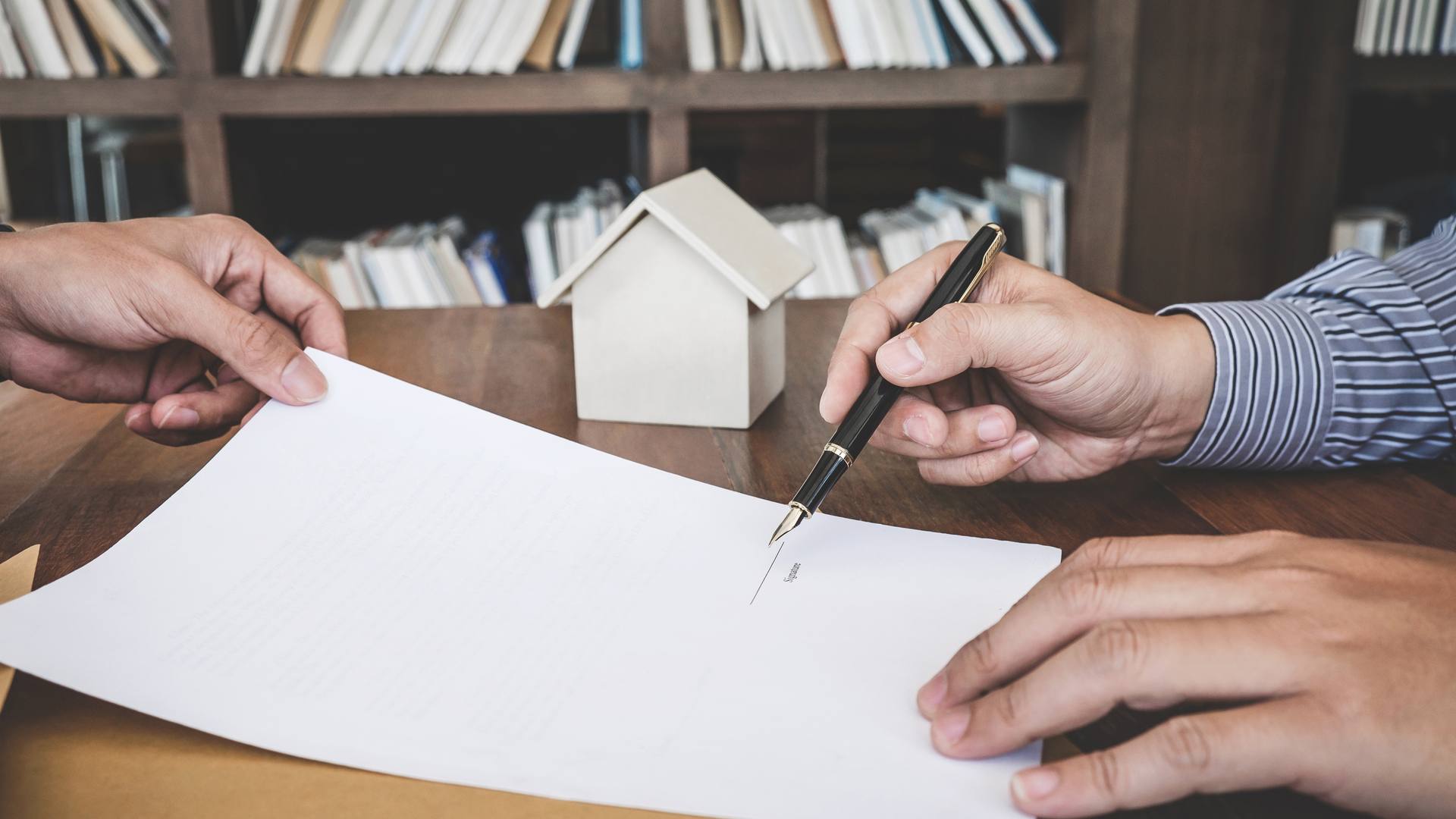 Una mano señala con una pluma la línea de firma de un contrato, junto a una casita de madera, en una mesa delante de una estantería con libros